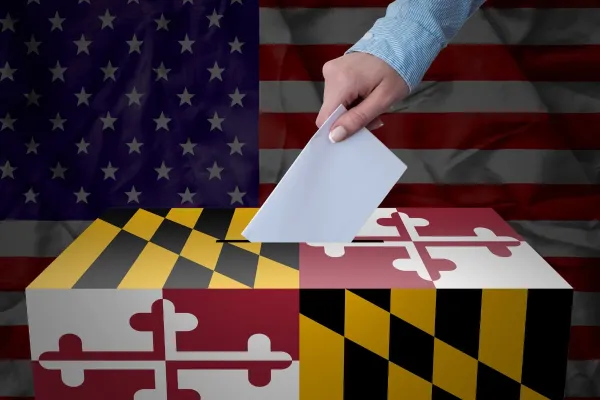 Woman's hand casts ballot in box wrapped in Maryland flag in front of an American flag.
