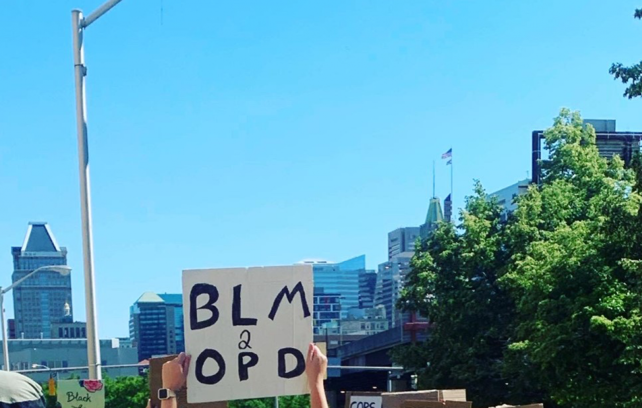 Baltimore skyline with a sign in the foreground saying "BLM 2 OPD"